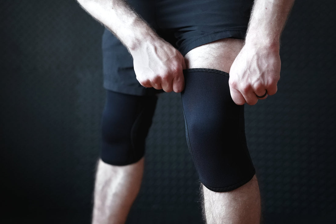 Bay Gear Co. black 7mm knee sleeves with embossed back logos next to a 50 ml liquid chalk bottle